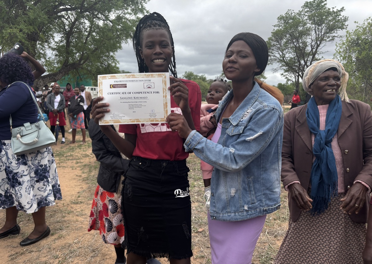 A young woman holds up her certificate of competence, standing outdoors beside two women.