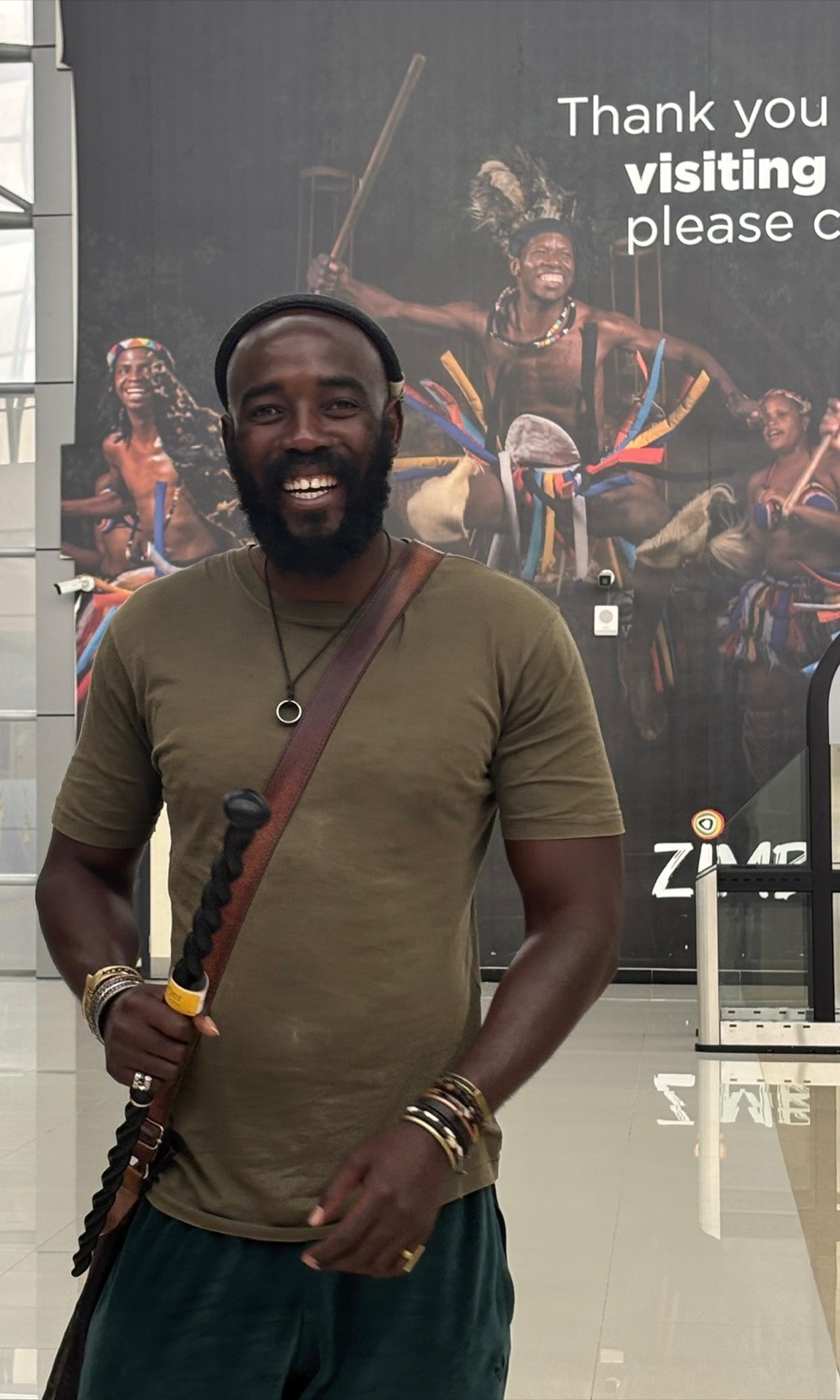 Chief Dakamela smiles, holding a carved walking stick, in front of a large poster of traditional dancers.