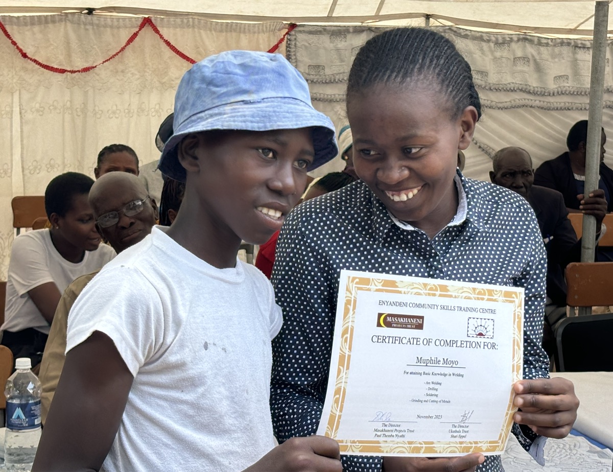 A young man in a bucket hat and a smiling young woman hold a certificate of completion from the Enyandeni Community Skills Training Centre.
