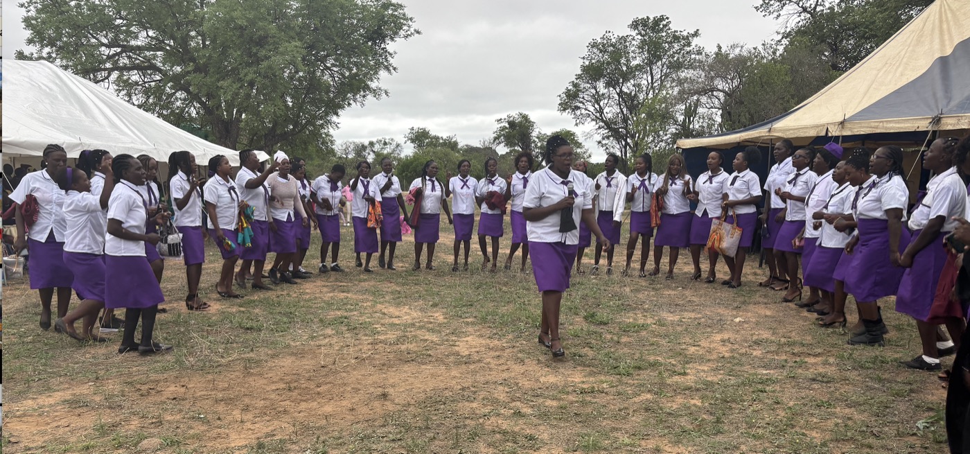 A large group of women and girls in purple skirts and white blouses gather on open ground between two marquees.