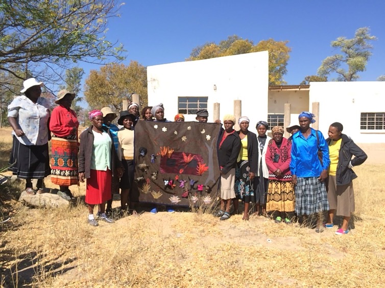 A group of women stand outside a building, holding up a large textile artwork they made together.