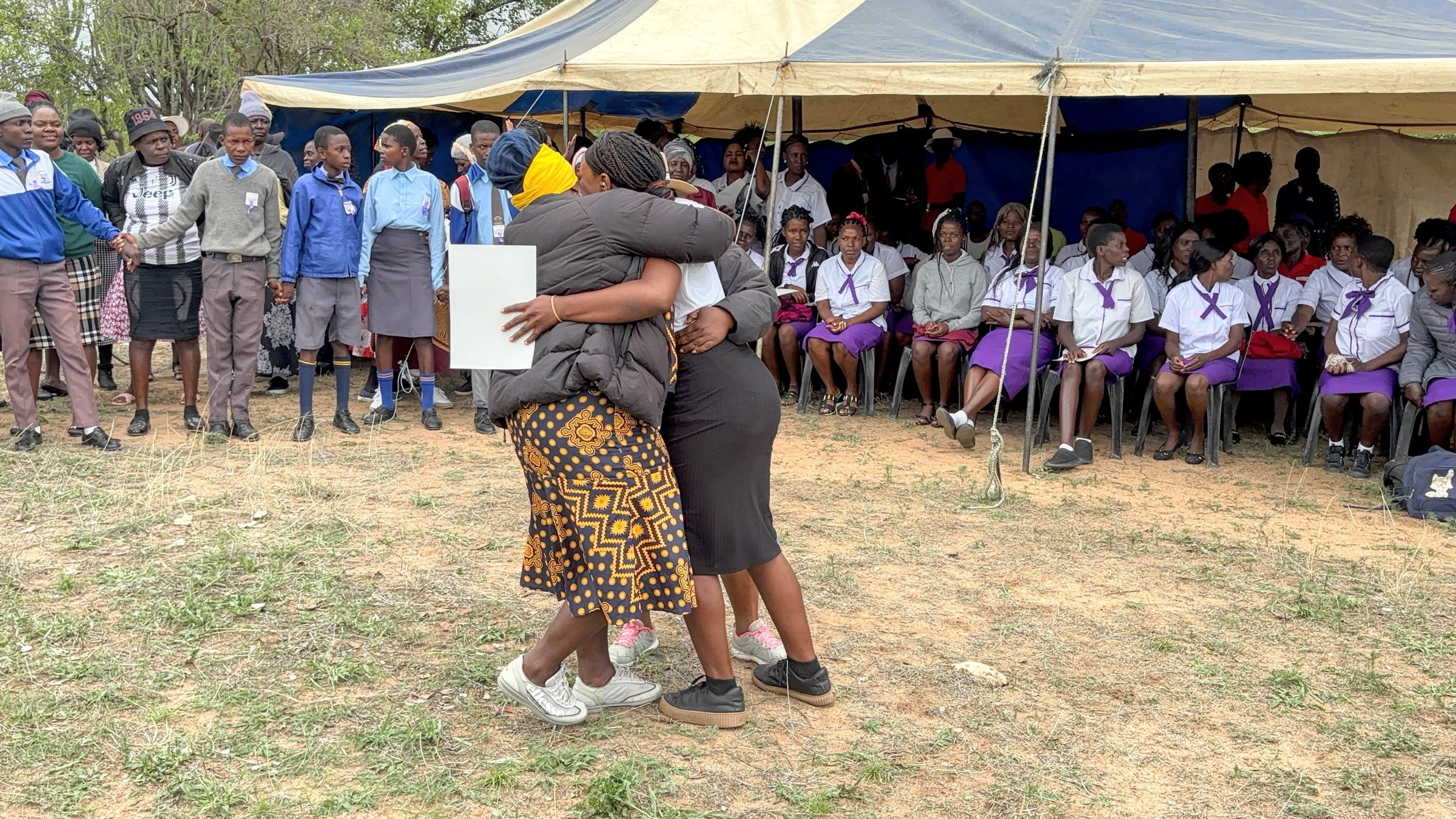 Two women embrace on an open field at an outdoor ceremony, while students in purple and white school uniforms look on from under a marquee.