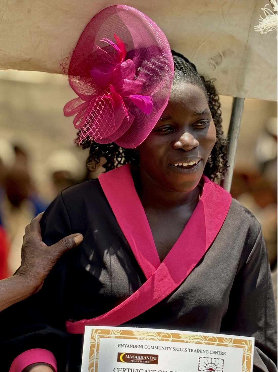 A smiling young woman in a pink fascinator holds a certificate from the Enyandeni Community Skills Training Centre.