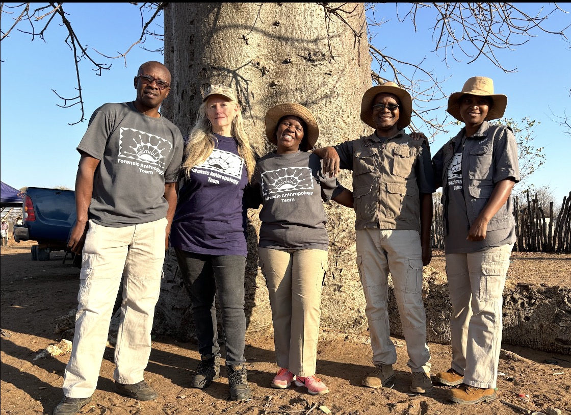 Five people in matching t-shirts and field clothes stand together in front of a large baobab tree.