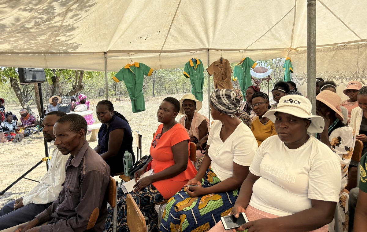 Community members, most of them women, sit together under a marquee during a meeting, with uniforms hanging from the tent behind them.